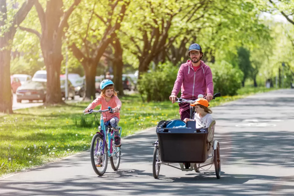 Father riding cargo bike with one child in cargo basket and one child riding bike alongside on sunny tree-lined path